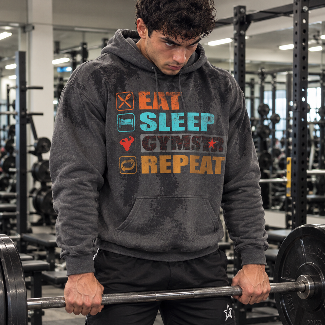 Luca Moretti wearing a dark heather GYMSTAR pullover hoodie with a colorful Eat Sleep GYMSTAR Repeat graphic and visible sweat marks, performing barbell shrugs in a gym.