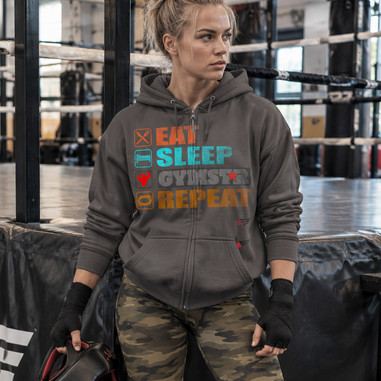 GYMSTAR model Emelie Johansson wearing a steel grey Eat Sleep GYMSTAR Repeat zip hoodie and camouflage leggings, holding boxing headgear beside a boxing ring.