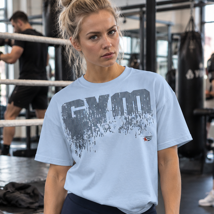 Emelie Johansson wearing a light blue GYMSTAR graphic T-shirt and dark navy leggings, posing with a water bottle in a boxing gym with athletes training in the background.