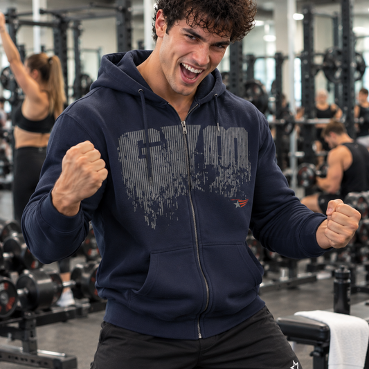 Luca Moretti celebrating a successful workout in a navy blue GYMSTAR full-zip hoodie, raising his fists in a bright gym with other athletes training in the background.